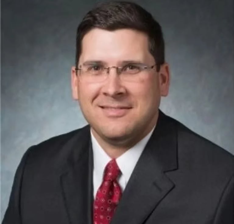 Professional headshot of a man wearing glasses, black blazer, and burgundy tie against a gray background